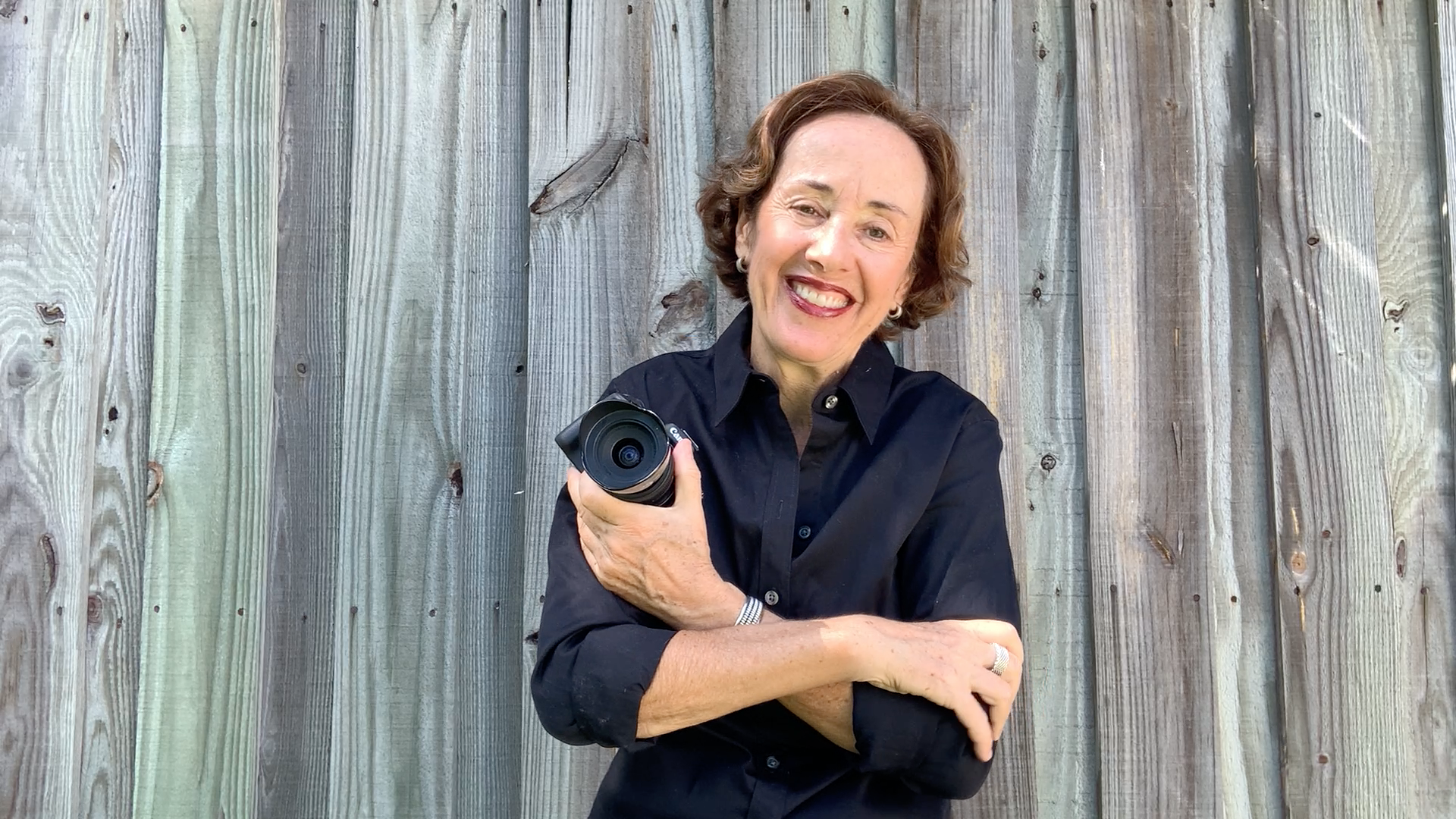 digital media producer Jana Panarites wearing black dress shirt, holding camera, with grey-colored fence in background